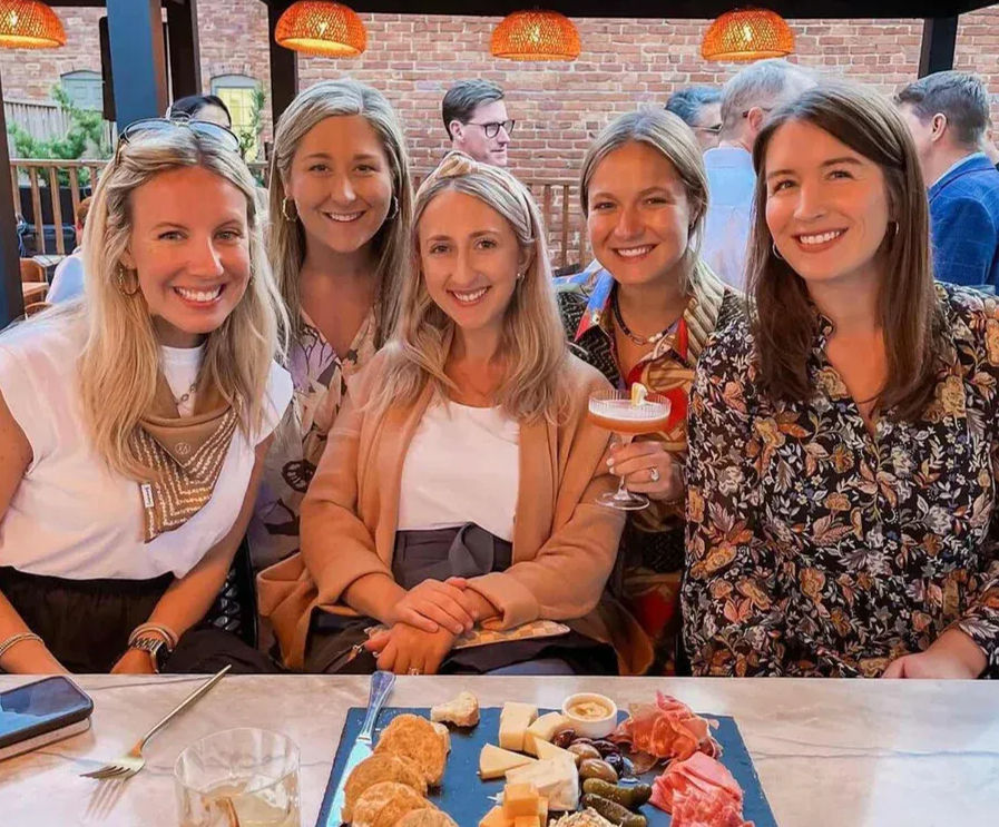 Five smiling friends gathered on an urban brick‑walled outdoor patio, sharing a charcuterie board and cocktails under warm pendant lights.
