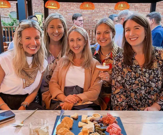 Five smiling friends gathered on an urban brick‑walled outdoor patio, sharing a charcuterie board and cocktails under warm pendant lights.