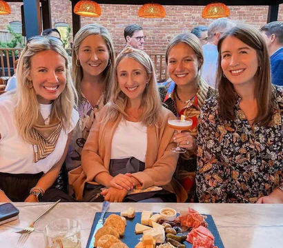 Five smiling friends gathered on an urban brick‑walled outdoor patio, sharing a charcuterie board and cocktails under warm pendant lights.