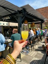 Hand holding a yellow mimosa in a champagne flute with a lipstick kiss at a sunny rooftop bar patio during outdoor brunch, patrons seated at the bar