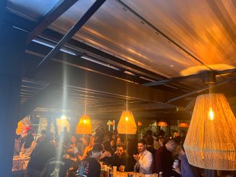 Crowded cozy bar interior at night with warm woven pendant lamps, exposed wooden beams and brick walls, groups of people socializing and drinking at the bar and tables.