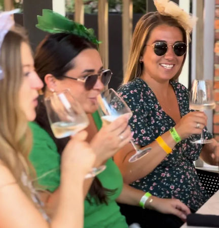 Three women on a sunny outdoor patio brunch, wearing sunglasses and festive headpieces, smiling and sipping white wine from stemmed glasses — one in green and another in a floral dress with event wristbands.