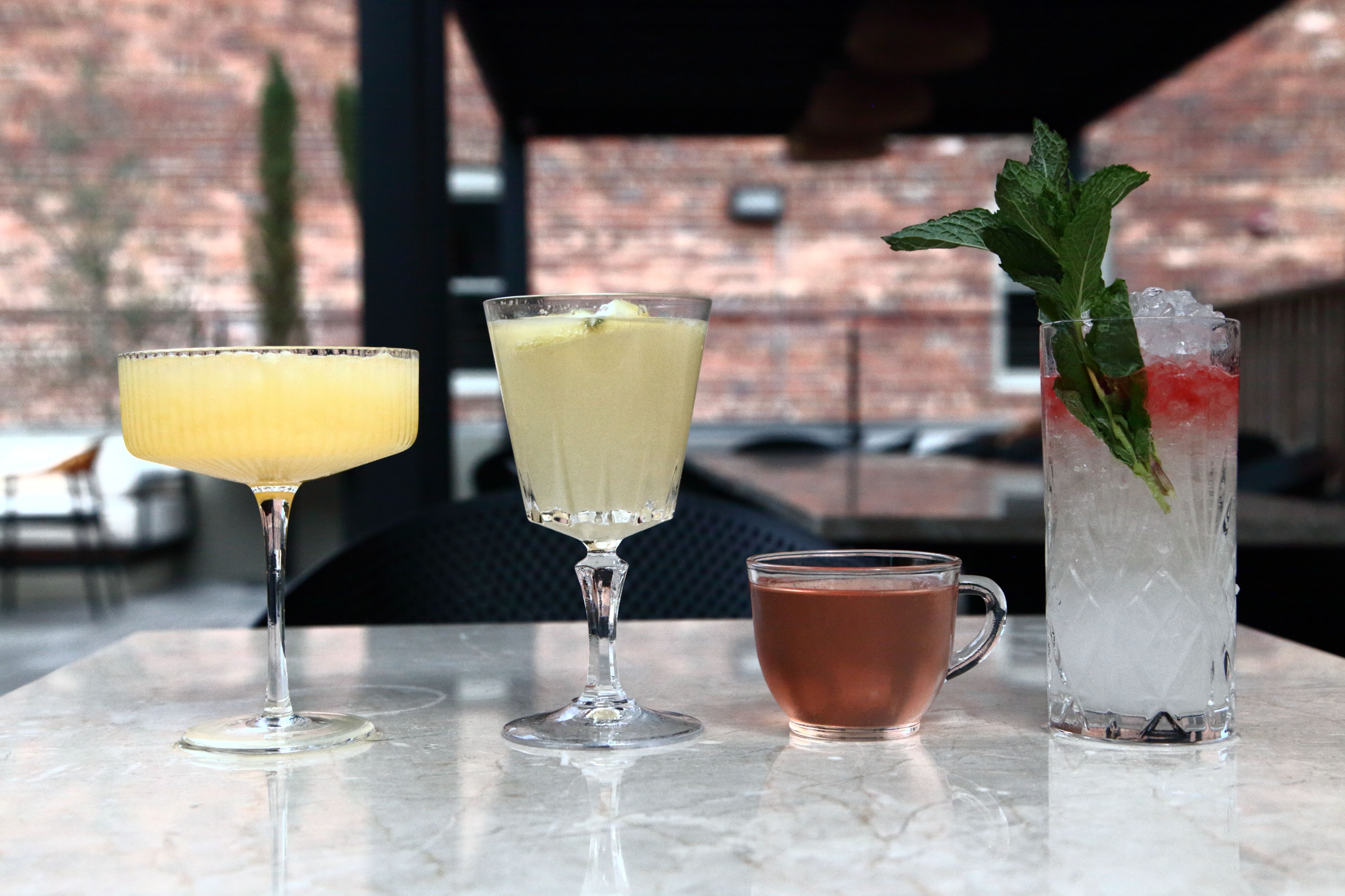 Four assorted drinks on a marble table at an outdoor patio: a chilled yellow coupe cocktail, a lemon-garnished stemmed drink, a small cup of amber tea, and a tall crushed-ice cocktail with mint and red garnish against a brick wall backdrop.