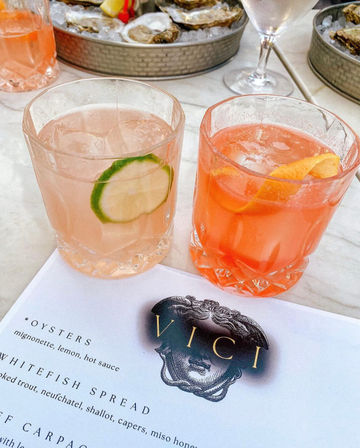Two pink cocktails in faceted tumblers — a pale drink with a lime wheel and a coral drink with an orange twist — on a marble table next to trays of iced oysters and a seafood menu at a coastal seafood restaurant.
