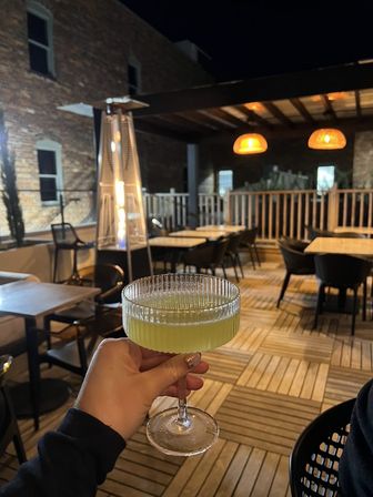 Cozy rooftop patio at night with a pale green cocktail in a ribbed coupe glass held above wooden decking, outdoor heater, rattan pendant lights and empty tables and chairs in the background.