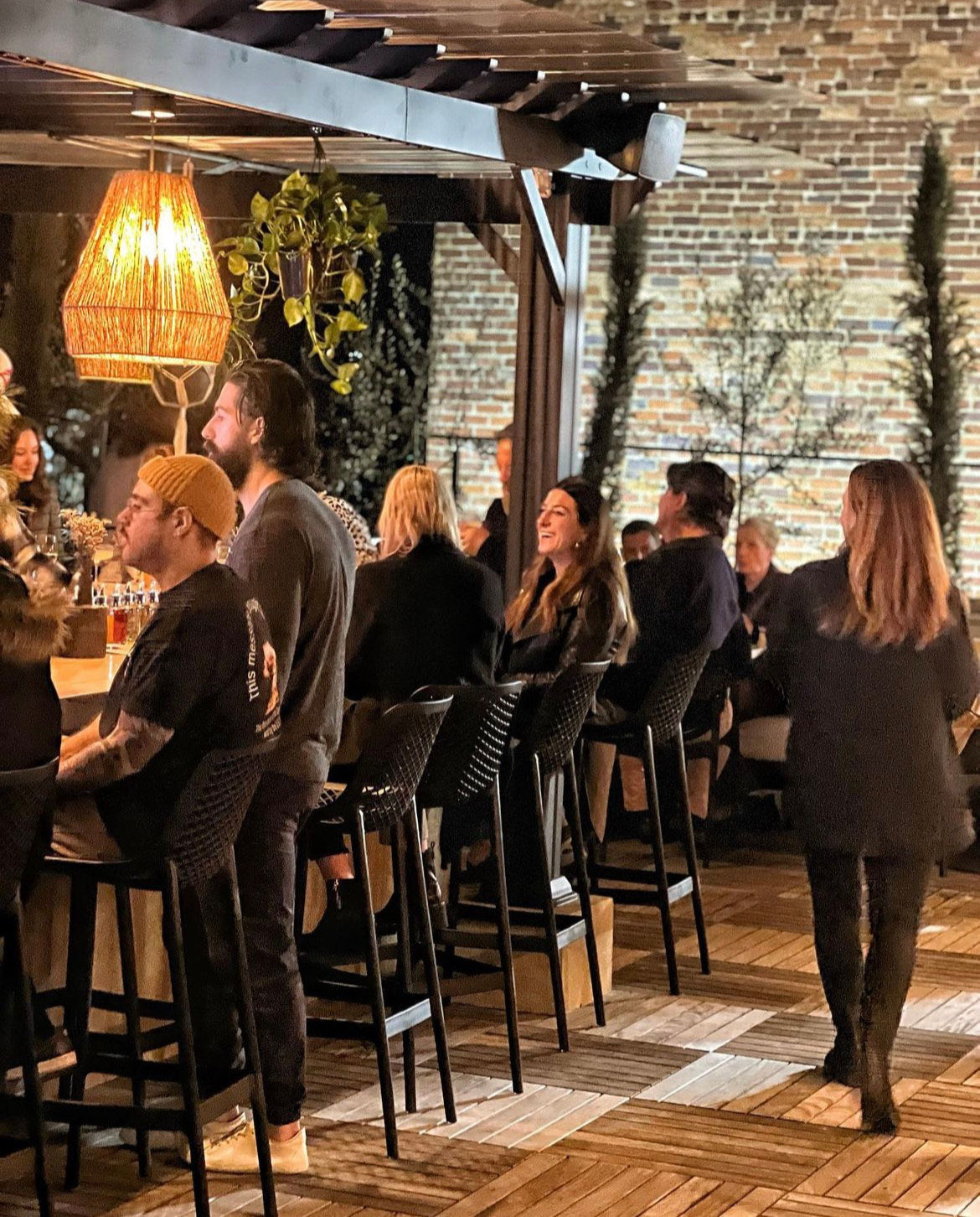 Evening rooftop patio bar scene with people seated on high stools at a wooden bar under a pergola, warm hanging lamp, hanging plants and exposed brick wall in the background.