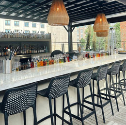 Sunlit rooftop outdoor bar with a long marble countertop, row of black woven bar stools, colorful bitters and liqueur bottles lined up, wicker pendant lights and city buildings in the background.