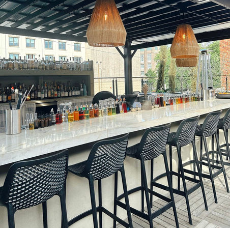 Sunlit rooftop outdoor bar with a long marble countertop, row of black woven bar stools, colorful bitters and liqueur bottles lined up, wicker pendant lights and city buildings in the background.