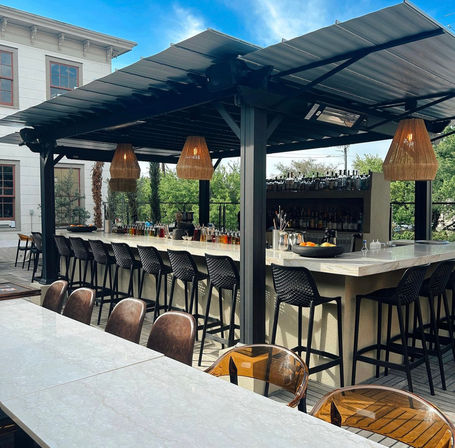 Sunlit outdoor rooftop bar patio with marble counters, a row of black high stools under a metal pergola, woven rattan pendant lights and a stocked back bar overlooking leafy trees.