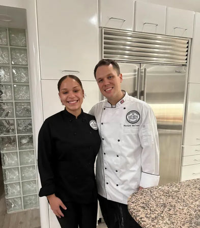 Two smiling chefs in black and white chef coats pose in a bright, modern kitchen with a stainless-steel refrigerator and granite countertop.