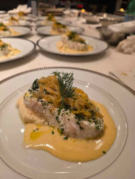 Plated white fish fillet on mashed potatoes with creamy hollandaise-style sauce, pickled onions and dill garnish on white china, multiple identical plates lined up in a restaurant kitchen