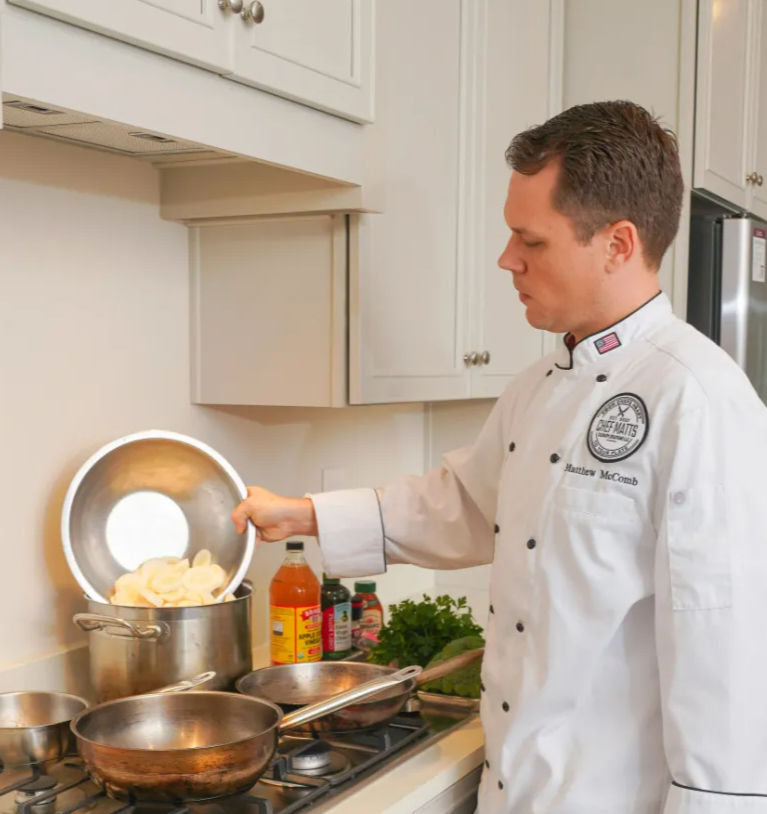 Chef in a white jacket pouring thinly sliced potatoes from a mixing bowl into a large pot on a home kitchen stovetop, with stainless pans and fresh herbs nearby.