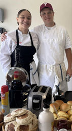 Two smiling chefs in aprons behind a busy breakfast station with a toaster, waffle press, baskets of English muffins and rolls, syrup bottle and jar of jam — cafe kitchen ready for morning service.