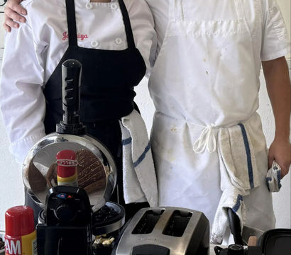 Two smiling chefs in aprons behind a busy breakfast station with a toaster, waffle press, baskets of English muffins and rolls, syrup bottle and jar of jam — cafe kitchen ready for morning service.