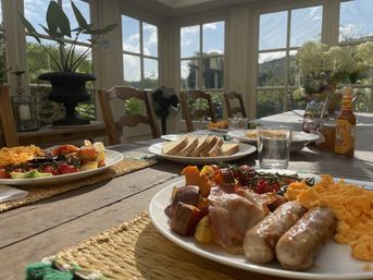 Sunlit farmhouse sunroom breakfast: plates of scrambled eggs, sausages, bacon and roasted cherry tomatoes with sliced bread, condiments and glasses on a rustic wooden table by large paned windows overlooking a green garden.