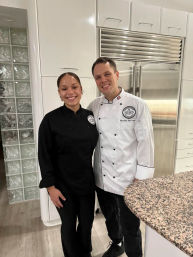 Two smiling chefs in black and white chef coats pose in a bright modern white kitchen with a stainless-steel refrigerator and speckled granite countertop.