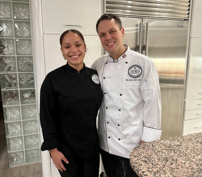 Two smiling chefs in black and white chef coats pose in a bright modern white kitchen with a stainless-steel refrigerator and speckled granite countertop.