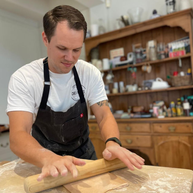 Home baker in white t-shirt and black apron rolling dough with a wooden rolling pin on a floured countertop in a cozy kitchen with a wooden hutch and shelves in the background.