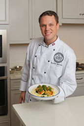 Smiling chef in a white coat presenting a plate of seared scallops on a creamy puree topped with herb gremolata in a bright modern kitchen