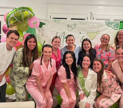 Smiling friends in colorful matching pajamas and robes pose indoors in front of lime-and-pink balloons and a cocktail-themed backdrop at a pajama party celebration.