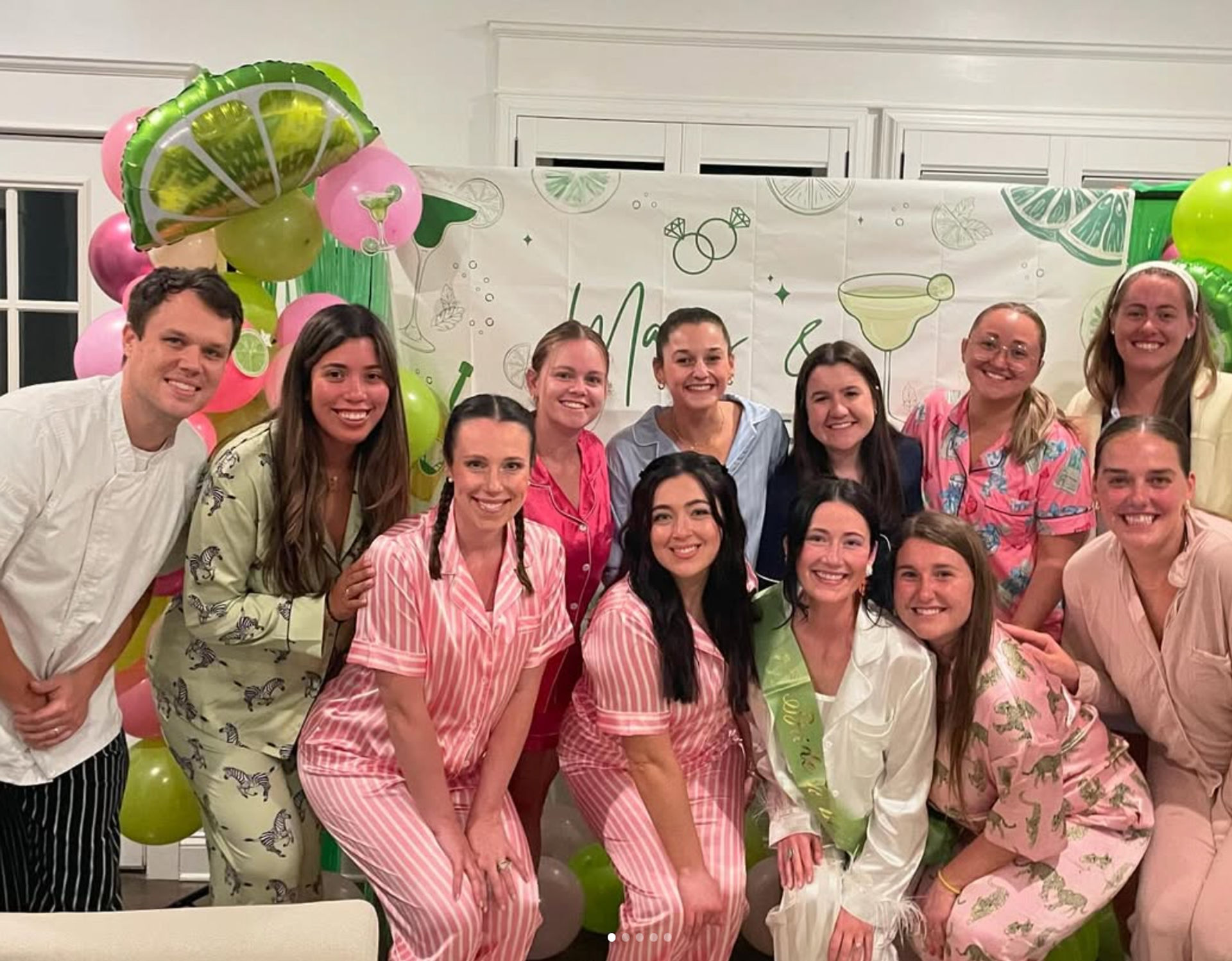Smiling friends in colorful matching pajamas and robes pose indoors in front of lime-and-pink balloons and a cocktail-themed backdrop at a pajama party celebration.