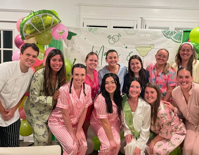 Smiling friends in colorful matching pajamas and robes pose indoors in front of lime-and-pink balloons and a cocktail-themed backdrop at a pajama party celebration.