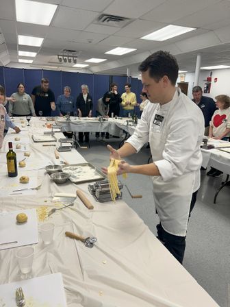 Instructor demonstrating fresh pasta-making with a hand-crank pasta machine, stretching golden fettuccine while a group of participants watches around long tables in a community cooking class.