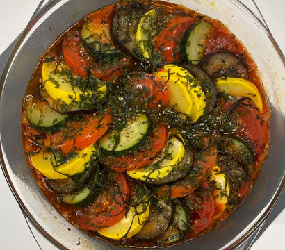 Vibrant ratatouille-style baked vegetables in a round glass dish — layered zucchini, yellow squash, eggplant and tomato slices in herbed tomato sauce.