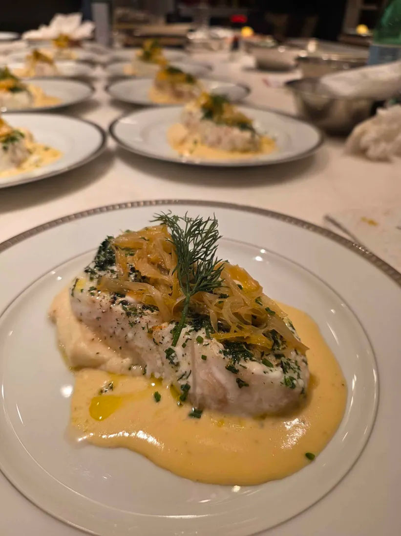 Close-up of a plated white fish fillet on a creamy yellow sauce over mashed potatoes, topped with caramelized onions, chopped chives and a dill sprig, with multiple similar plates lined up in a restaurant kitchen.