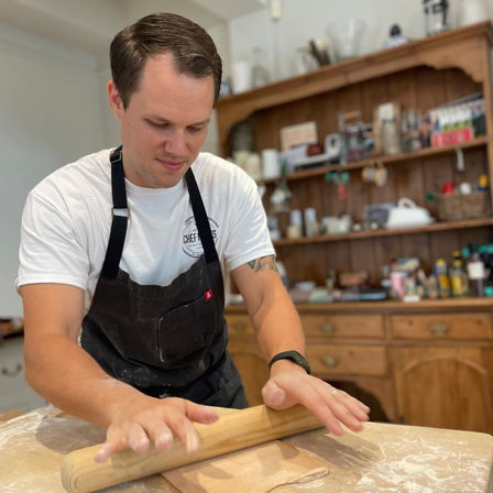 Home baker in a black apron rolling dough with a wooden rolling pin on a floured countertop in a cozy, rustic kitchen with a wooden hutch and jars on open shelves.