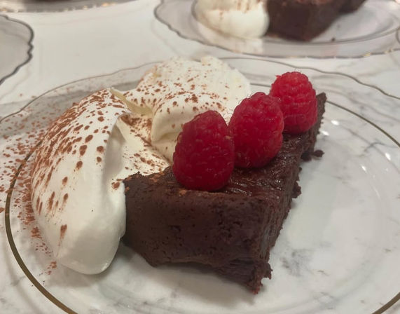 Decadent chocolate tart slice topped with three fresh raspberries, served with cocoa-dusted whipped cream on a decorative dessert plate.