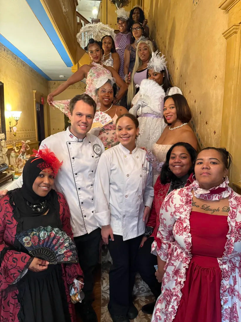 Group portrait on an ornate gold-painted staircase: two chefs in white coats surrounded by people wearing Victorian-style dresses, fascinators, fans and parasols, festive tea-party vibe.