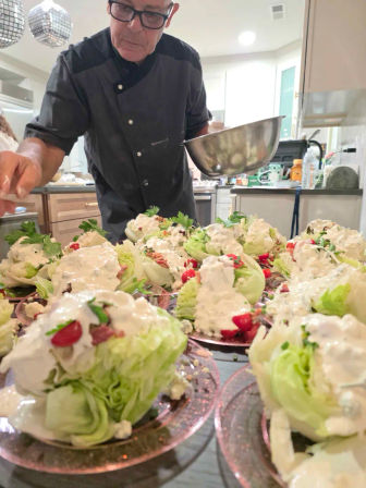 Close-up of a cook plating iceberg wedge salads topped with creamy dressing, cherry tomatoes, bacon bits and parsley on pink plates in a bright kitchen