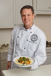 Smiling chef in a white jacket holds a white-rimmed bowl of seared scallops over creamy polenta, topped with vibrant green herb sauce, posed in a bright modern kitchen.