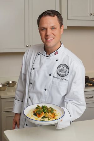 Smiling chef in a white jacket holds a white-rimmed bowl of seared scallops over creamy polenta, topped with vibrant green herb sauce, posed in a bright modern kitchen.