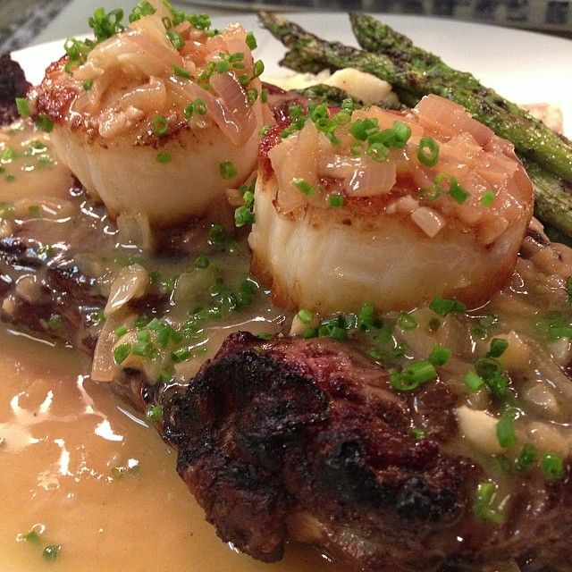 Close-up of seared scallops topped with caramelized shallots and chopped chives in a savory pan sauce, plated as surf-and-turf with charred steak and grilled asparagus.