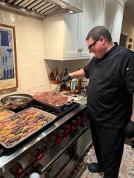 Smiling cook in black uniform and gloves arranging seasoned steaks on a pan over a gas range in a bright home kitchen, with a tray of multicolored fingerling potatoes and pans on the stovetop.