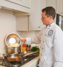 Chef in a white jacket pouring sliced potatoes from a metal bowl into a large stockpot on a gas stove, with pans, bottles and fresh herbs on a bright home kitchen counter.
