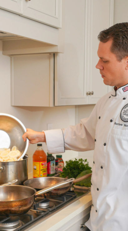 Chef in a white jacket pouring sliced potatoes from a metal bowl into a large stockpot on a gas stove, with pans, bottles and fresh herbs on a bright home kitchen counter.