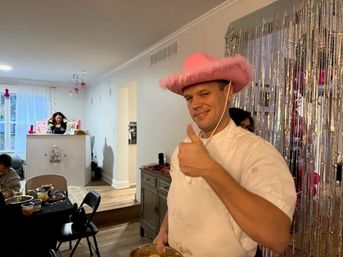 Man in a white shirt wearing a pink fuzzy cowboy hat gives a thumbs-up at a home party with silver fringe backdrop, pink decorations and a table of food.