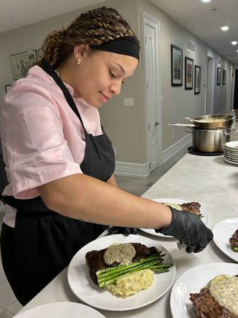 Cook plating steak topped with herb butter, grilled asparagus and mashed potatoes on white plates at a modern kitchen counter