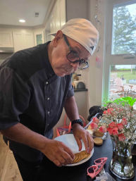 Man in a backwards white cap and glasses arranging crackers on a plate on a kitchen island next to a vase of pink roses and pink sunglasses, with a windowed backyard view