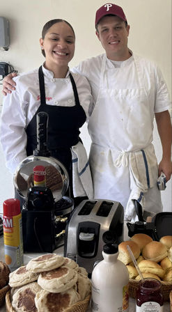 Two smiling chefs in white uniforms behind a breakfast counter with baskets of English muffins and rolls, a toaster and waffle iron, a bottle of maple syrup and jar of jam — cozy local cafe breakfast spread.