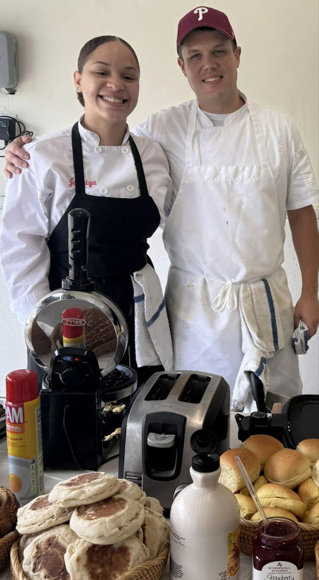Two smiling chefs in white uniforms behind a breakfast counter with baskets of English muffins and rolls, a toaster and waffle iron, a bottle of maple syrup and jar of jam — cozy local cafe breakfast spread.