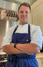 Smiling chef in a white jacket and navy apron with arms crossed in front of a stainless-steel range and hood in a bright home kitchen, ready to cook.