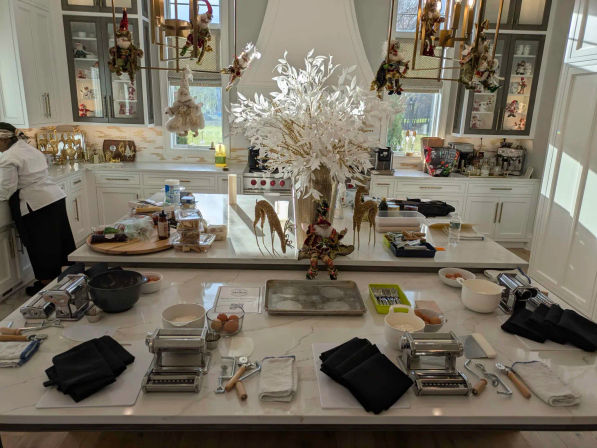 Bright white gourmet kitchen with marble island set up for a festive pasta-making class — multiple manual pasta machines, bowls with eggs, utensils, trays, black napkins and a large white floral centerpiece.