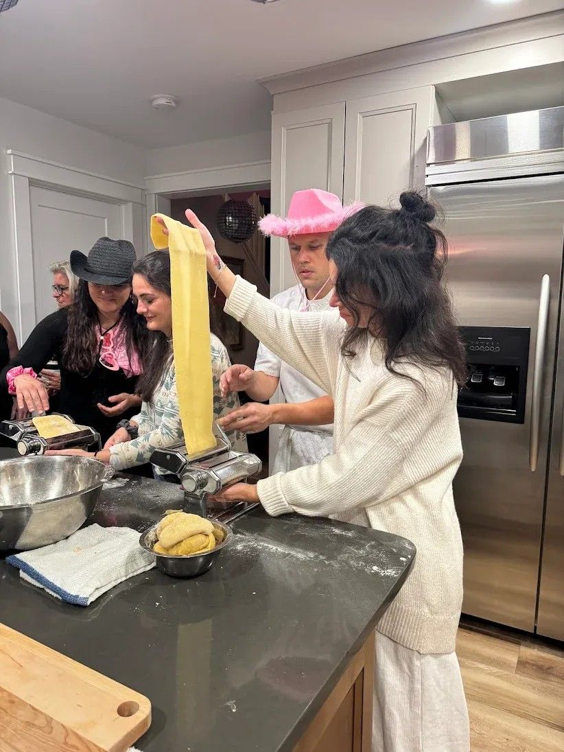 Group making homemade pasta in a home kitchen — hand-crank pasta machine rolling a long sheet of dough, mixing bowls and playful hats.