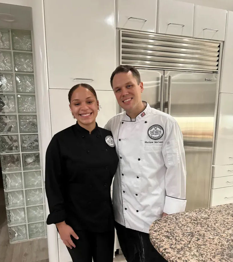 Two smiling chefs posing in a modern white kitchen — one wearing a black chef coat and one in a white chef coat — standing by a stainless-steel refrigerator and granite countertop.