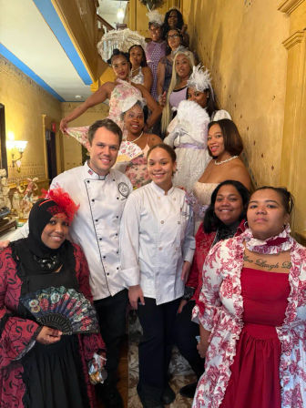 Group portrait of people in Victorian-style dresses, fascinators and fans with two chefs posed on an ornate staircase, holding parasols for a themed tea party in a historic parlor