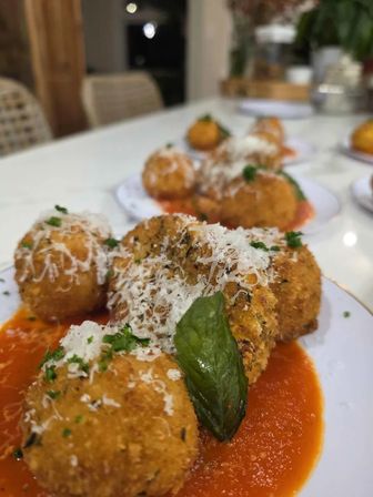 Close-up of golden fried arancini (risotto balls) on white plates over marinara sauce, sprinkled with grated Parmesan and parsley and garnished with a basil leaf — Italian appetizer in a modern dining setting.